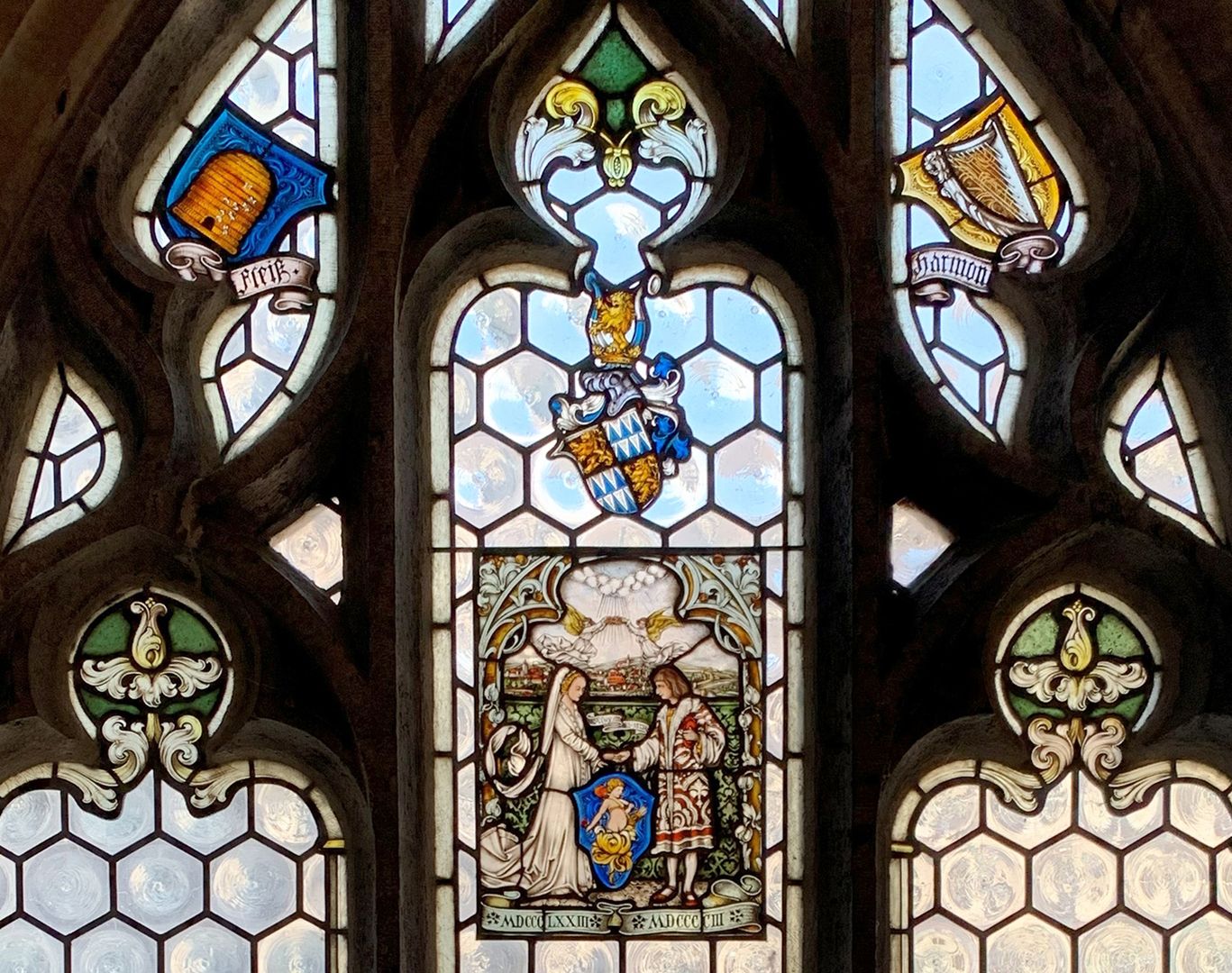 Fleiß und Harmonie oberes Fenster, Spitzbogen mit Maßwerk / oben: Links ein Bienenkorb mit dem Wort "Fleiß" unterschrieben ist. Rechts eine gelb hinterlegte Harfe "mit der Unterschrift „Harmonie“