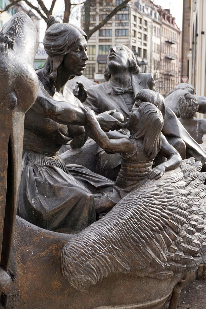 Ehekarussell / Hans-Sachs-Brunnen "Pelikanwagen" / "Die in ein faltenreiches blaues, abendkleid ähnliches Gewand gekleidet Mutter füttert die beiden Kinder". (J.W.)