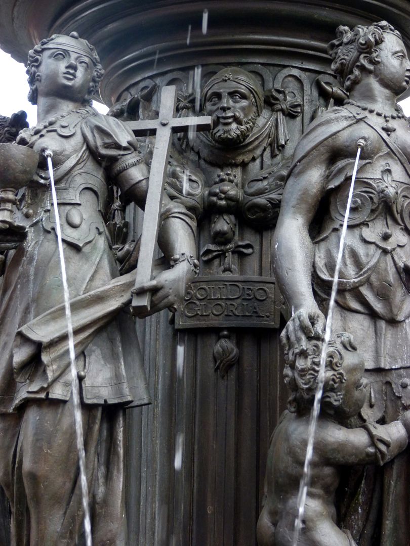 Tugendbrunnen Zwischen den Tugenden Glaube und Liebe auf der Brunnensäule eine Tafel mit der Inschrift: SOLI DEO GLORIA = Allein Gott zur Ehr